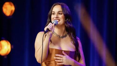 Female singer with long, dark wavy hair, wearing a strapless brown top and a gold-toned necklace, holding a microphone and smiling.
